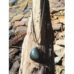 Pendentif obsidienne œil céleste en goutte sur cordon noir, suspendu à un tronc de bois flotté avec galets et eau scintillante en arrière-plan, pierre naturelle associée à l’ancrage, à la présence à soi et à la sérénité.