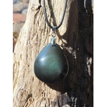 Pendentif obsidienne œil céleste en goutte aux reflets verts sur cordon noir, suspendu à un tronc de bois flotté au bord de l’eau, pierre naturelle liée à la clarté, à l’ancrage et à la protection subtile.