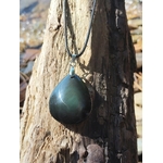 Pendentif obsidienne œil céleste en goutte sur cordon noir, suspendu à un tronc de bois au bord de l’eau, pierre naturelle associée à la protection, à la lucidité et à la stabilité intérieure.
