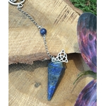 Pendule en lapis-lazuli bleu présenté sur du bois avec chaîne argentée, perle assortie et pierre décorative violette, dans une ambiance naturelle propice à l’intuition.