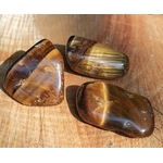 Trois pierres roulées œil de tigre aux reflets dorés et bruns sur fond bois chaleureux, pierres naturelles associées à la stabilité, à l’aplomb et à la présence à soi.
