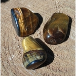 Trois pierres roulées œil de tigre aux reflets bruns et dorés posées sur une surface sableuse, pierres naturelles associées à l’enracinement, à la maîtrise de soi et à l’assurance.