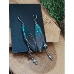 Boucles d’oreilles fée création Magic Gaïa présentées sur support noir, posées sur bois avec mousse verte, ailes bleu-vert irisées et pampilles fines, dans une ambiance féerique évoquant douceur et légèreté.