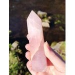 Main tenant une pointe de quartz rose facettée en extérieur, fond nature flou, pour inviter à la tendresse, apaiser les émotions et encourager l’amour de soi.