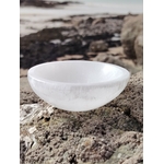 Bol chantant en sélénite 10 cm posé sur un rocher granitique face à la mer en Bretagne, galets et ciel voilé en décor, pour une ambiance apaisante propice à la clarté intérieure et au recentrage.