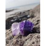 Deux améthystes brutes violettes sur rocher breton, mer floue en arrière-plan et lumière douce, pour apaiser les émotions, clarifier l’esprit et protéger.