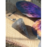 Pendule en pierre de lune noire posé sur une surface en bois clair, près de fleurs violettes et pêche, dans une ambiance lumineuse et mystique évoquant intuition, ancrage et protection intérieure.