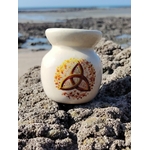 Brûleur en céramique blanc orné d’une triquetra dorée, posé sur les rochers en Bretagne face à la mer, dans une lumière douce, évoquant apaisement, harmonie et ancrage