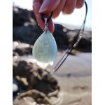 Main tenant un pendentif en aigue-marine au-dessus des rochers et de l’eau en Bretagne, bijou en pierre naturelle lié à la fluidité, à la sensibilité paisible et au recentrage.