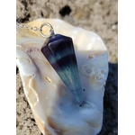 Pendule en fluorite posé dans un coquillage clair sur fond sableux, outil de divination à l’ambiance douce évoquant intuition, clarté mentale et recentrage.