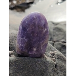 Lépidolite forme libre violette posée sur roche sombre au bord de l’eau, pierre naturelle associée au calme des pensées, à l’équilibre émotionnel et au recentrage.