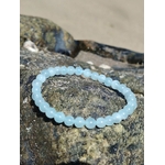 Bracelet Aigue-Marine 6 mm posé sur rochers en bord de mer, perles bleu lagon lumineuses, sérénité intérieure et communication douce.