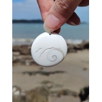 Pendentif œil de Sainte Lucie blanc présenté à la main devant la plage et l’horizon marin, coquillage naturel à spirale beige, paix intérieure et chance.
