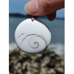 Pendentif œil de Sainte Lucie rond blanc tenu entre les doigts, spirale beige visible sur fond marin, coquillage porte-bonheur lié à la chance et au calme intérieur.