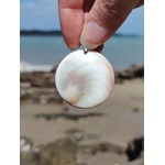 Face nacrée d’un pendentif œil de Sainte Lucie rond blanc tenue entre les doigts, sur fond de plage et horizon marin, sérénité et énergie apaisante.
