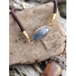 Zoom sur bracelet médaillon ovale en labradorite facettée posé sur pierre avec gland en décor naturel, cordon brun et monture dorée, intuition et stabilité émotionnelle.