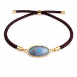 Bracelet réglable en labradorite facettée sur fond clair uni, médaillon ovale gris bleu à monture dorée et cordon brun, élégance discrète et énergie protectrice.