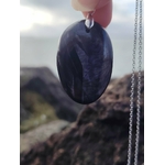 Pendentif Hyperstène tenu à la main devant le littoral breton, pierre noire ovale à reflets argentés, protection symbolique et recentrage