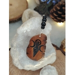 Pendentif de rétroviseur face Gaïa posé sur cristal de roche brut, bois pyrogravé et tourmaline noire dans un décor naturel lumineux.