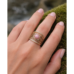 Bague dorée multi-rangs portée au doigt sur décor de mousse, pierre rose ronde et monture travaillée au style bohème élégant.