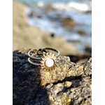 Bague Soleil cabochon nacré photographiée sur roche côtière, bijou doré lumineux avec mer floue en arrière-plan.