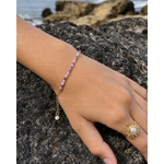 Main posée sur un rocher en bord de mer, bracelet fin en améthyste facettée sur chaîne dorée avec breloque et bague soleil dorée à perle blanche, dans une ambiance lumineuse évoquant sérénité et intuition.