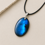 Pendentif ovale en Labradorite bleue sur fond beige, labradorescence vive, cordon noir et finition argentée pour élégance et recentrage.