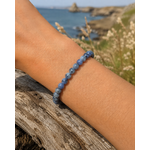 Bracelet Tanzanite perles 6-7 mm porté au poignet sur bois flotté, mer et falaises de Bretagne en arrière-plan, intuition, expression et accomplissement.