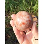 Sphère en Pierre de Soleil naturelle tenue en main, finition polie, reflets cuivrés scintillants, fond herbe flou — symbole de joie et d’optimisme.