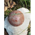 Sphère en Pierre de Soleil naturelle (héliolite) posée sur un amas de quartz blanc, nuances pêche-abricot avec inclusions scintillantes, brin d’herbe au premier plan — symbole de joie, d’optimisme et d’énergie positive.
