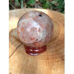 Sphère en Pierre de Soleil naturelle (héliolite) sur support bois, posée sur grand rondin, reflets nacrés orangés, fond végétal flou — symbole de joie, d’optimisme et d’énergie positive.