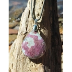 Pendentif en tourmaline rose naturelle, forme ronde polie sur cordon tressé, posé sur bois flotté en extérieur, pierre naturelle associée à la tendresse, au réconfort et à l’équilibre émotionnel.