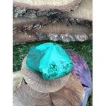 Pierre roulée de malachite, veines sombres visibles, décor nature (bûches, herbes) — calme intérieur, volonté, clarification du mental, chakra coeur