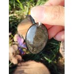 Pendentif ovale en obsidienne dorée avec éclats dorés, présenté dans la main en décor nature, pierre naturelle reliée au discernement, à la confiance calme et à la présence à soi.