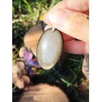 Pendentif obsidienne dorée tenu dans la main sur fond d’herbe et de fleurs, bijou en pierre naturelle associé à l’enracinement, à la solidité intérieure et à la sérénité.