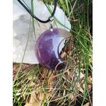 Pendentif lune en améthyste dans l’herbe contre un amas de quartz, cordon noir visible, bijou naturel associé à la paix intérieure, à l’intuition et au réconfort émotionnel.