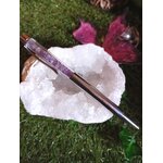 Stylo bille rempli d’éclats d’améthyste posé sur une grande druse de cristal blanc avec pétales de rose sur l’herbe, accessoire d’écriture en pierres naturelles pour créativité et apaisement du mental.