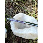 Stylo bille transparent rempli d’éclats d’améthyste, posé sur une grande druse de quartz blanc dans l’herbe, accessoire d’écriture en pierres naturelles pour créativité et calme intérieur.