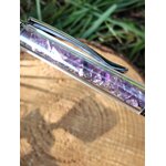 Gros plan sur un stylo bille transparent rempli d’éclats d’améthyste violette, posé en diagonale sur une rondelle de bois avec de l’herbe en arrière-plan, accessoire d’écriture en pierres naturelles favorisant créativité et sérénité.