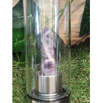 Gros plan sur la pointe d’améthyste naturelle dans la bouteille d’eau en verre énergisante, base inox posée sur l’herbe, accessoire de lithothérapie pour le bien-être.
