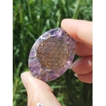 Petit galet orgonite améthyste avec fleur de vie dorée tenu entre les doigts devant un fond de nature, galet énergétique de lithothérapie pour harmonisation des énergies, protection, apaisement mental et émotionnel