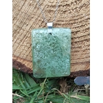 Pendentif rectangulaire en fluorite verte sur chaîne argentée, posé devant un rondin de bois et de l’herbe, pierre naturelle liée à la légèreté intérieure, à la justesse émotionnelle et au recentrage.