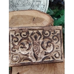 Boîte à bijoux en bois sculpté motif déesse, posée sur une tranche de bois, avec une grande rondelle de bois, un disque décoratif gris et du feuillage vert en arrière-plan.