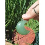 Pendentif lune en aventurine verte tenu entre les doigts devant l’herbe, bijou en pierre naturelle évoquant le lâcher-prise, la tendresse et l’équilibre affectif.