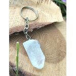 Porte-clés / bijou de sac en cristal de roche taillé en pointe facettée, avec chaîne et anneau métal, suspendu devant une tranche de bois clair avec une petite tige verte en décor naturel