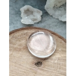 Galet cristal de roche poli posé sur tranche de bois avec quartz brut en arrière-plan, pierre roulée translucide associée à la pureté, à l’amplification et à l’harmonie intérieure.