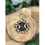 Porte-encens rond en bois motif Triple Lune, posé sur une tranche de bois avec feuillage vert en arrière-plan.