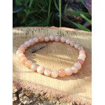 Bracelet pierre de soleil 6 mm rose orangé lumineux en perles rondes posé sur un support en bois, avec feuillage sombre en arrière-plan, évoquant l’enthousiasme, la vitalité et la confiance intérieure