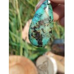 Gros plan sur pendentif turquoise africaine goutte bleu-vert marbrée de noir, tenu entre les doigts devant feuillage, jaspe africain évoquant la lucidité, le recentrage et la confiance tranquille.