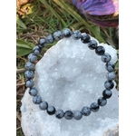 Bracelet obsidienne neige 6 mm posé sur galet clair, perles noir profond et gris nuage, feuillage en arrière-plan, protection douce, ancrage, calme intérieur, pensées plus nettes.